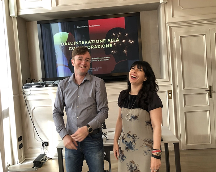 Giacomo Bosio e Cristiana Melis sorridenti davanti al banner dell’evento 'Dall’interazione alla collaborazione' presso Copernico Milano Centrale.
